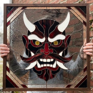 Pode incluir: Um painel de vitral quadrado numa moldura de madeira escura com um design de máscara Hannya. A máscara é composta por peças de vidro vermelho escuro, preto e branco com olhos amarelos, sobre um fundo de vidro texturizado transparente com bordas geométricas.