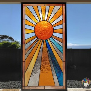 May include: A stained glass window depicting a sunburst design. The sun is a large orange circle with rays in orange, yellow, blue, and silver. The window is framed in black and set against a blue sky. The design is reminiscent of Art Deco.