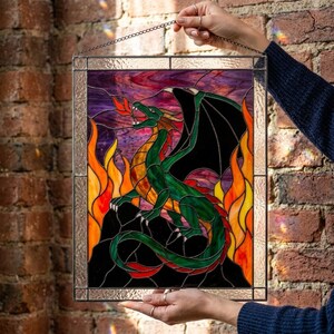 Op de afbeelding: Een rechthoekig glas-in-loodpaneel met een groene draak met zwarte vleugels op een zwarte rotsformatie. De draak is omringd door oranje en gele vlammen tegen een paarse lucht. Het paneel is omlijst met een dunne metalen rand.
