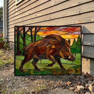 Puede incluir: Un panel rectangular de vidrieras que muestra un jabalí marrón en un entorno forestal. El fondo presenta colinas verdes, siluetas de árboles oscuros y un cielo vibrante al atardecer con bandas horizontales de vidrio naranja, amarillo y morado.