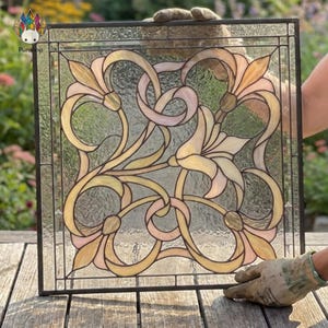 Könnte beinhalten: Quadratische Glasmalerei mit floralem Design in Gelb-, Rosa- und Cremetönen. Das Glas ist schwarz umrandet. Das Design zeigt stilisierte Blumen und wirbelnde Linien. Gehalten von behandschuhten Händen.