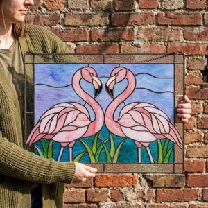 Puede incluir: Un panel rectangular de vidrieras que muestra dos flamencos rosas frente a frente con los cuellos curvados. Las aves están situadas sobre un cielo azul claro y hierba verde, todo ello enmarcado en un marco de metal oscuro con una cadena.