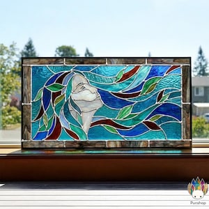 May include: Stained glass panel depicting a woman's profile with flowing hair. The artwork uses shades of blue, green, and red, with a white face. The panel is framed in a dark wood-toned border and sits on a wooden surface.