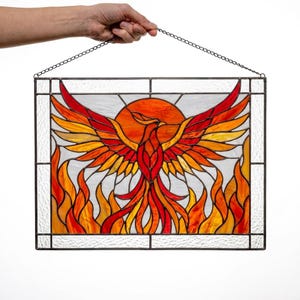 Op de afbeelding: Een rechthoekig glas-in-loodpaneel met een feniks met gespreide vleugels in tinten rood, oranje en geel. De vogel is gecentreerd voor een cirkelvormige oranje zon, omringd door gestileerde vlammen aan de basis, alles omlijst door helder glas.