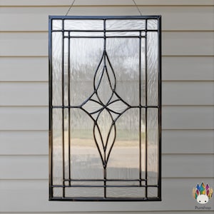 Puede incluir: Panel de ventana rectangular de vidrieras con marco de metal negro. El vidrio transparente presenta un patrón texturizado y un diseño geométrico negro central. El panel está suspendido por una cadena, un elemento decorativo.