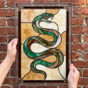 Pode incluir: Um painel retangular de vitral apresentando uma cobra enrolada com a boca aberta. A cobra é feita de vidro verde e castanho matizado, sobre um fundo de peças de vidro creme e bege texturizadas, tudo emoldurado em metal escuro.