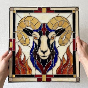 Könnte beinhalten: Ein quadratisches Buntglasfenster mit einem Widderkopf und großen, geschwungenen Hörnern. Das Design besteht aus cremefarbenen, bernsteinfarbenen, blauen, roten und schwarzen Glasteilen, die durch dunkle Metallstege verbunden sind, vor einem cremefarbenen Hintergrund.