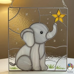 Op de afbeelding: Gebrandschilderd glaspaneel met een grijze olifant die naar een gele ster reikt. De olifant staat tegen een gouden en witte achtergrond met een maan- en sterrenontwerp. Het paneel is omlijst met metaal en heeft een ophangketting.