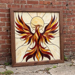 Può includere: Un pannello quadrato in vetro colorato raffigurante una fenice con le ali spiegate nei toni del rosso, arancione e giallo, su uno sfondo color crema con un motivo solare circolare, il tutto racchiuso in una cornice di legno marrone scuro.