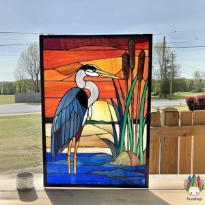 Könnte beinhalten: Buntglasscheibe mit einem blauen Reiher im Wasser, mit Schilf und einem Sonnenuntergang im Hintergrund. Das Kunstwerk ist schwarz gerahmt und im Freien ausgestellt. Der Reiher hat einen langen gelben Schnabel und ist von grünen Schilf umgeben.