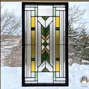 Puede incluir: Una ventana de vitrales con un diseño geométrico en tonos de verde, amarillo y blanco. La ventana está enmarcada en negro y se coloca sobre un fondo nevado.