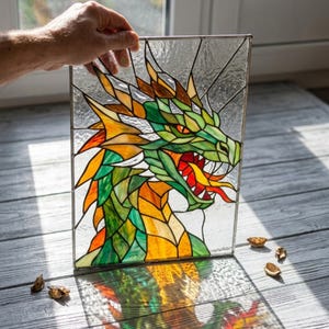 Op de afbeelding: Gebrandschilderd glaspaneel met een drakenkop. De draak is afgebeeld met groene, oranje en gele schubben, rode ogen en een open mond. Het kunstwerk is omlijst met een metalen rand en wordt door een hand vastgehouden.