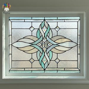 May include: A rectangular stained glass window panel featuring a central symmetrical design of interlocking teal and cream-colored glass pieces. The pattern is surrounded by textured, clear glass panes held together by dark metal lead lines.