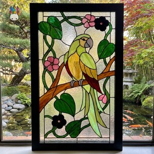 Peut inclure: Un panneau en vitrail rectangulaire dans un cadre noir représentant un perroquet vert et jaune perché sur une branche brune. Le motif comprend des feuilles vertes, des fleurs roses et des fleurs noires sur un fond en verre texturé translucide.