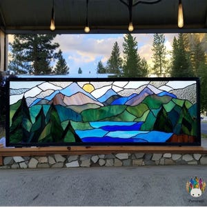 Pode incluir: Um painel de vitral retratando uma paisagem montanhosa com um lago, árvores e um sol poente. A obra de arte apresenta tons de azul, verde e castanho, com uma moldura preta. O design lembra uma vista panorâmica.