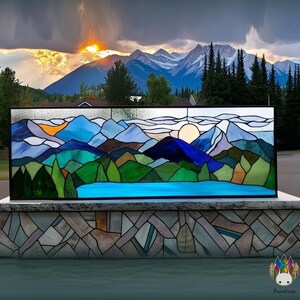 May include: Stained glass panel depicting a mountain landscape with a lake, trees, and a setting sun. The artwork features shades of blue, green, orange, and brown, framed in black. The background shows a mountain range and a dramatic sunset.