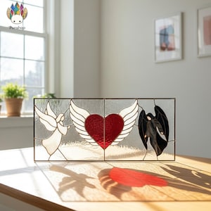 Puede incluir: Panel de vidrieras rectangular con tres diseños: un ángel blanco, un corazón rojo con alas blancas y un ángel negro. El panel está sobre una superficie de madera. El diseño está iluminado por luz natural.