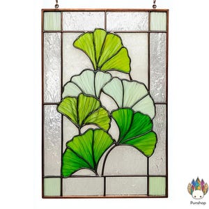 Puede incluir: Panel de vidrieras con un diseño de hojas de ginkgo. Las hojas son de varios tonos de verde, con un aspecto texturizado. El panel está enmarcado en metal marrón oscuro y tiene una cadena para colgar. El fondo es blanco esmerilado.