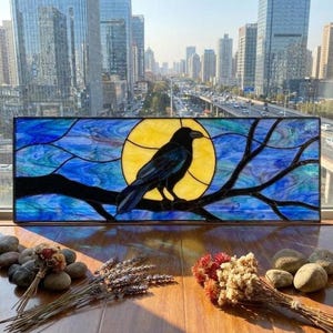 Puede incluir: Un panel rectangular de vidriera que muestra un cuervo negro posado sobre una rama oscura frente a una gran luna amarilla circular. El fondo consiste en segmentos de vidrio azul y turquesa que crean un efecto de cielo nocturno con líneas.