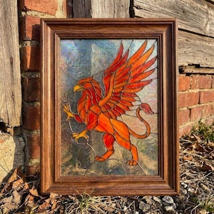 Può includere: Un pannello rettangolare in vetro colorato raffigurante un grifone arancione e rosso brillante con le ali spiegate, racchiuso in una cornice di legno marrone scuro. Il vetro ha una finitura testurizzata e iridescente, e il grifone è raffigurato di profilo.