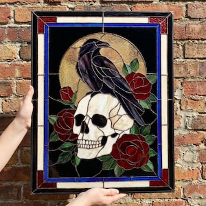 Può includere: Un pannello rettangolare in vetro colorato raffigurante un corvo nero appollaiato su un teschio umano color crema. Il design include rose rosso scuro con foglie verdi su sfondo nero, il tutto incorniciato da un bordo in vetro blu, bianco e rosso.