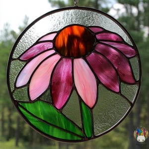 May include: A stained glass flower suncatcher with a circular design. The flower features pink and purple petals, a brown center, and green leaves. The glass is framed in a dark metal and hangs from a chain.