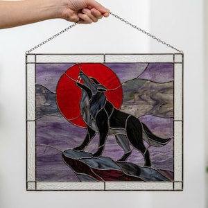 May include: A square stained glass panel featuring a black wolf howling at a large red moon. The background consists of swirling purple and white glass pieces. The entire piece is framed in metal and hangs from a thin silver-colored metal chain.