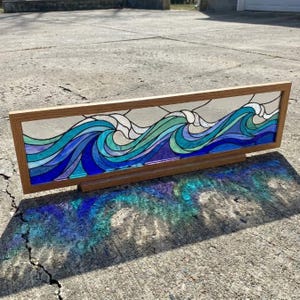 May include: A rectangular stained glass panel framed in light wood, featuring a continuous design of stylized ocean waves in shades of deep blue, turquoise, and seafoam green, with white accents representing crests, set within a simple wooden frame.