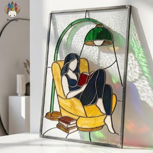 May include: A rectangular stained glass panel featuring a person sitting in a yellow armchair reading a red book under a green floor lamp. The design includes a stack of books on the floor, all encased in a dark metal frame with textured glass.