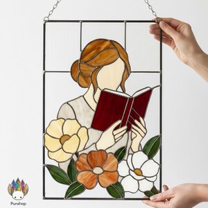 May include: Stained glass panel featuring a woman reading a book, with flowers at the bottom. The woman has brown hair and is wearing a light-colored top. The book is red. The flowers are yellow, orange, and white, with green leaves. The panel is framed in black.