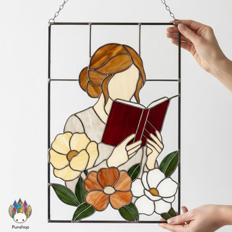 May include: Stained glass panel featuring a woman reading a book, with flowers at the bottom. The woman has brown hair and is wearing a light-colored top. The book is red. The flowers are yellow, orange, and white, with green leaves. The panel is framed in black.