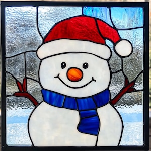 May include: Stained glass panel featuring a smiling snowman wearing a red Santa hat and a blue scarf. The snowman has a carrot nose and twig arms. The background is a light blue and white winter scene.