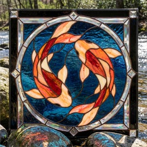 Può includere: Pannello in vetro colorato con due pesci koi in un design circolare. I pesci sono arancioni e rossi su uno sfondo blu, incorniciati da un bordo nero con dettagli in vetro trasparente. Il pannello è quadrato e decorativo.