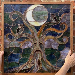 Pode incluir: Um painel de vitral quadrado numa moldura de madeira com uma árvore central com um rosto humano, rodeada por ramos em espiral. Duas corujas estão pousadas nos ramos sob uma grande lua crescente pálida contra um céu azul escuro.