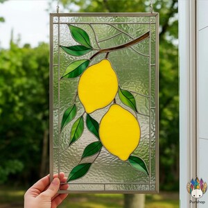 May include: Stained glass panel featuring two bright yellow lemons hanging from a brown branch with green leaves. The panel is framed in a silver-colored metal and is designed to hang. The background is a textured, clear glass.