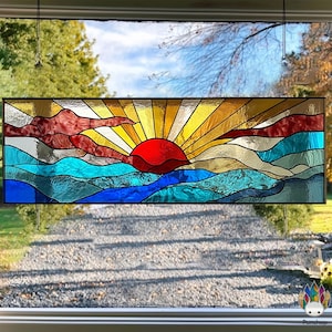 May include: A rectangular stained glass panel showing a sunset scene. The sun is a red circle, with golden rays. The sea is depicted with blue and turquoise waves, and the sky has red and orange clouds. The panel is framed in black.