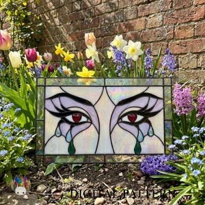 May include: A rectangular stained glass panel featuring a stylized design of two eyes with long black lashes, red heart-shaped pupils, and blue and green teardrops. The glass has an iridescent finish, and the text DIGITAL PATTERN is visible.