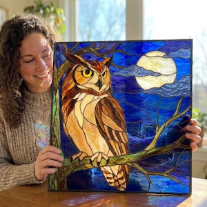 May include: A square stained glass panel featuring a brown and cream-colored owl perched on a tree branch. The background depicts a dark blue night sky with a large, circular, pale yellow moon and wispy clouds. The entire scene is framed in black.
