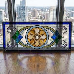 May include: A rectangular stained glass panel featuring a central yellow floral motif within a circular frame. The design includes green and blue diamond shapes, light purple curved accents, and clear glass jewels set against a dark blue border.