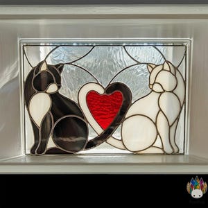 May include: Stained glass panel featuring two cats, one black and white, the other white. A red heart is between them. The panel is set in a white frame, with a textured glass background.