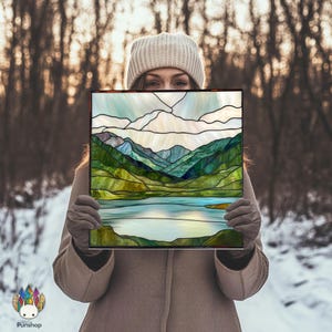 Peut inclure: Un panneau de vitrail carré représentant un paysage de montagne et de lac. L'œuvre d'art présente des nuances de vert, de bleu et de blanc. Le panneau est tenu devant une personne portant un manteau et un bonnet d'hiver.