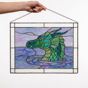 May include: A rectangular stained glass panel featuring a green dragon head emerging from purple and blue water. The dragon has iridescent scales and a sharp profile. The piece is framed in white textured glass and hangs from a metal chain.