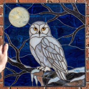 Puede incluir: Un panel de vidrieras cuadrado que muestra un búho blanco posado en una rama oscura contra un cielo nocturno azul profundo. Una gran luna llena de color amarillo pálido es visible en la esquina superior izquierda, rodeada de ramas desnudas.