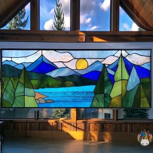 以下が含まれることがあります： 湖、木々、夕日を描いた山岳風景のステンドグラスパネル。 青、緑、黄色の色合いで、黒いフレームが特徴です。 画像は窓のアート作品です。