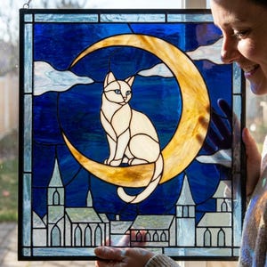 Op de afbeelding: Gebrandschilderd glaspaneel met een witte kat op een gouden maan tegen een blauwe achtergrond. Het ontwerp bevat een stadsgezicht en wolken. Het paneel is omlijst met zwarte lijnen.