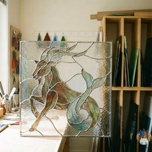 Puede incluir: Panel de vidriera cuadrado con un diseño del signo zodiacal de Capricornio. La obra de arte representa un híbrido de cabra y pez en tonos marrones, verdes y azules, sobre un fondo transparente. El panel está enmarcado con un borde plateado, mostrando un estilo artístico único.