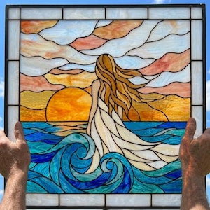 May include: A square stained glass panel depicting a person with long flowing hair standing in ocean waves at sunset. The scene features a bright orange sun, deep blue and turquoise water, and a sky filled with soft peach and white clouds.