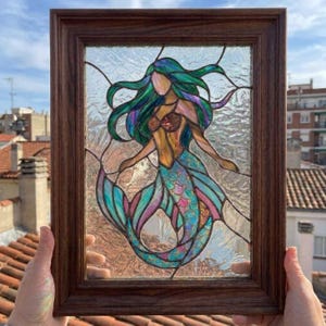 May include: A stained glass panel featuring a mermaid with flowing teal and purple hair, a tan bodice, and a shimmering iridescent tail. The artwork is encased in a dark brown wooden frame with a textured, clear glass background.
