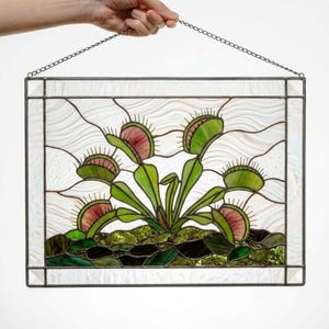 May include: A rectangular stained glass panel featuring a Venus flytrap plant with green leaves and pink-toned traps with spiky edges. The plant is set against a textured white background with a dark green base, all framed in metal with a hanging chain.
