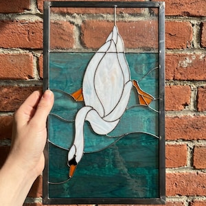 Peut inclure: Un panneau rectangulaire en vitrail représentant un cygne blanc aux pattes orange nageant dans une eau bleu sarcelle profonde. Le cygne est vu de dessus avec la tête immergée, entouré de lignes de verre ondulées, dans un cadre en métal.